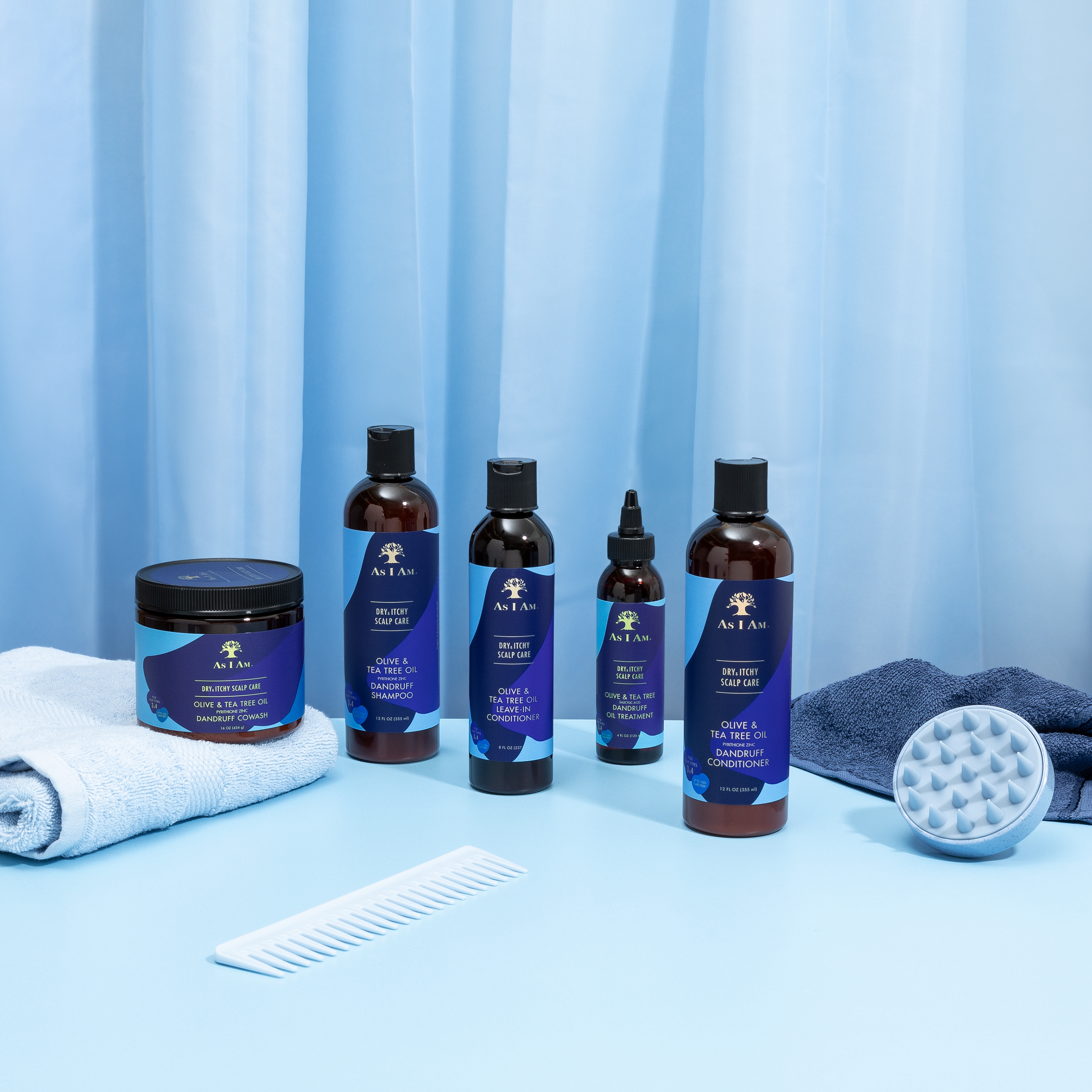 Produktbillede af As I Am Dry & Itchy Scalp Care serien, der inkluderer conditioner, oliebehandling og andre produkter, præsenteret med en kam, en massagebørste og håndklæder på en lyseblå baggrund.