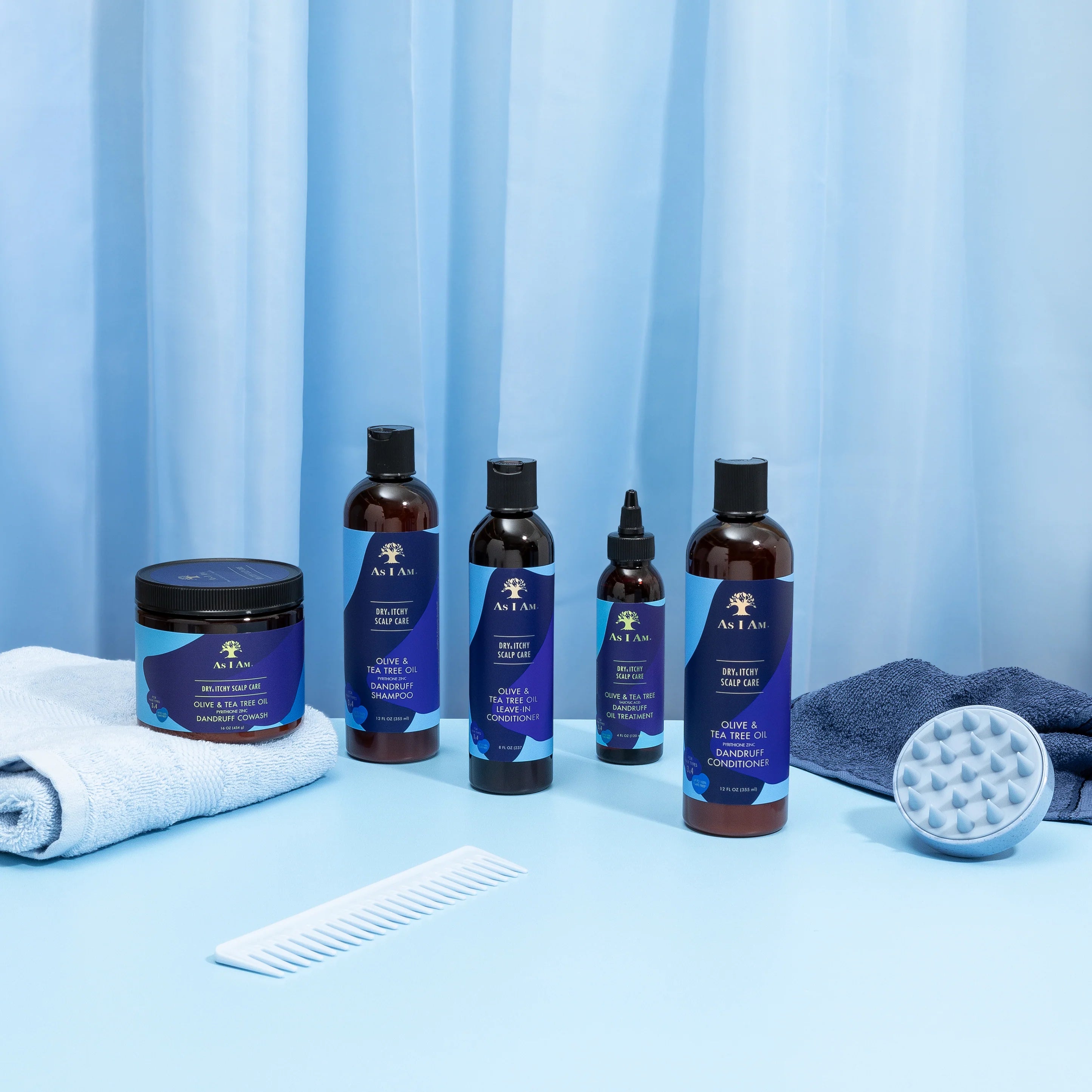 Forskellige produkter fra As I Am Dry & Itchy Scalp Care-serien er arrangeret på en lyseblå overflade med en hvid kam, en hvid vaskeklud og en grå hovedbundsbørste.
