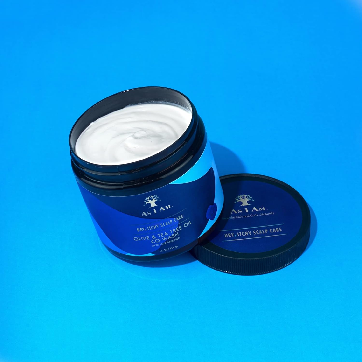 En åben beholder med As I Am Dry & Itchy Scalp Care CoWash står på en blå baggrund. Den hvide CoWash er synlig i beholderen, og låget ligger ved siden af. 