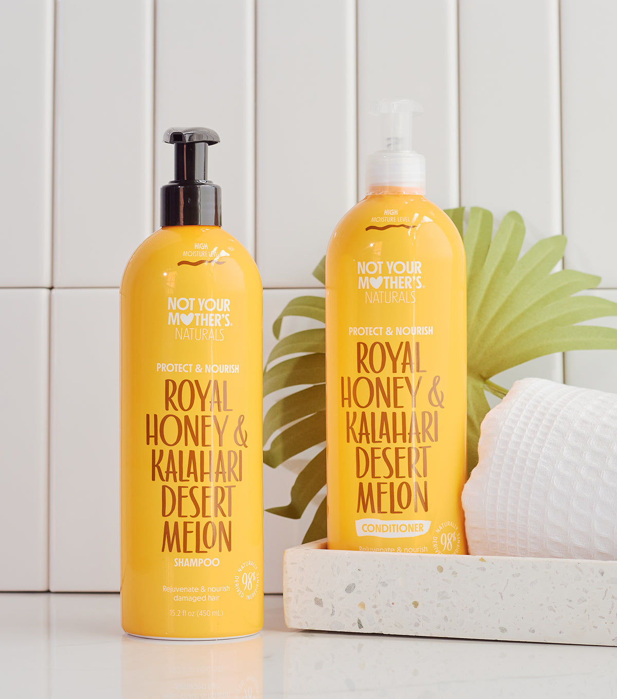 To gule flasker: Not Your Mother's Naturals Royal Honey & Kalahari Desert Melon shampoo (til venstre) og conditioner (til højre), stående på en hvid overflade foran en hvid flisevæg.
