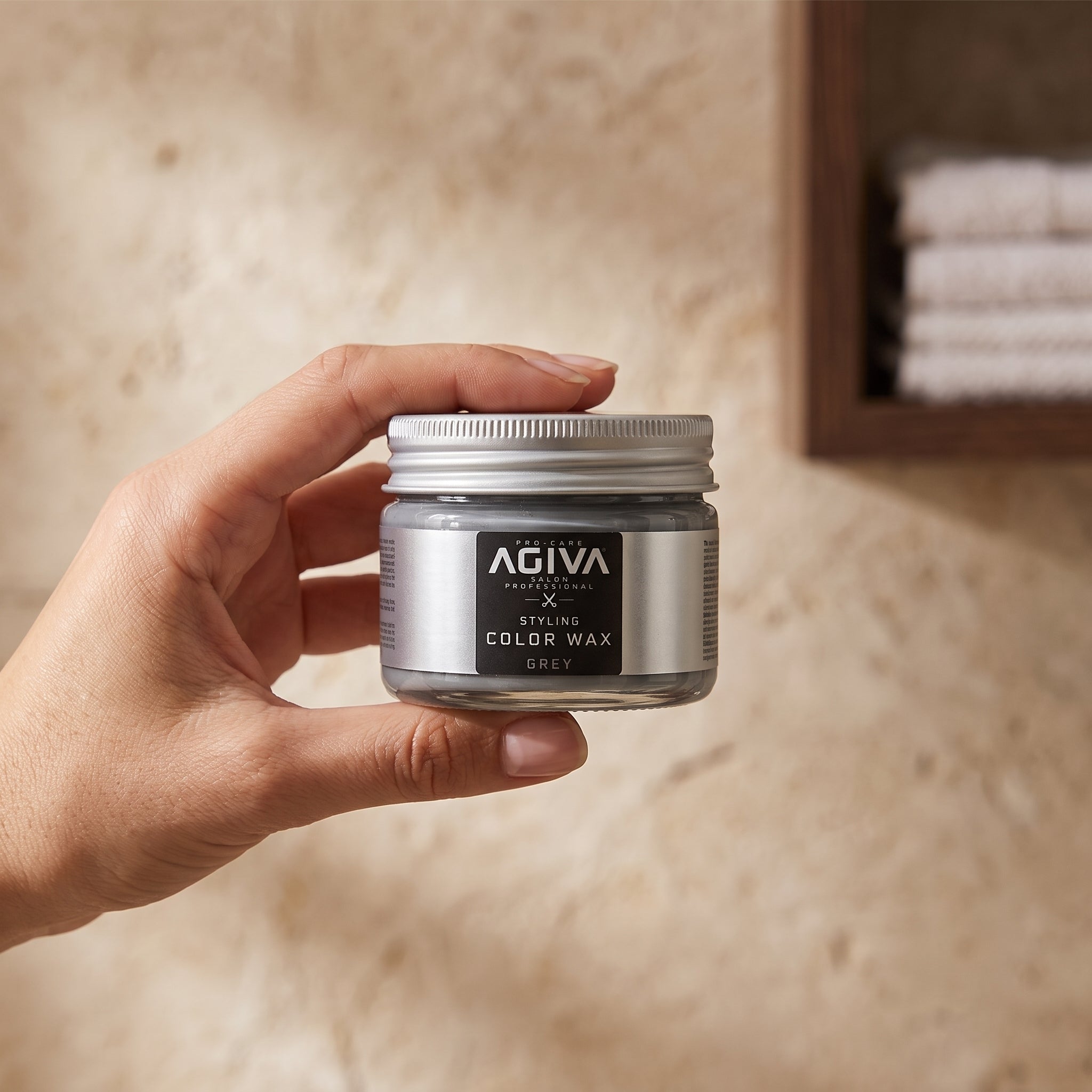 Hånd holder AGIVA Pro-Care Styling Color Wax i grå farve, glas med sølvlåg mod sandfarvet baggrund.
