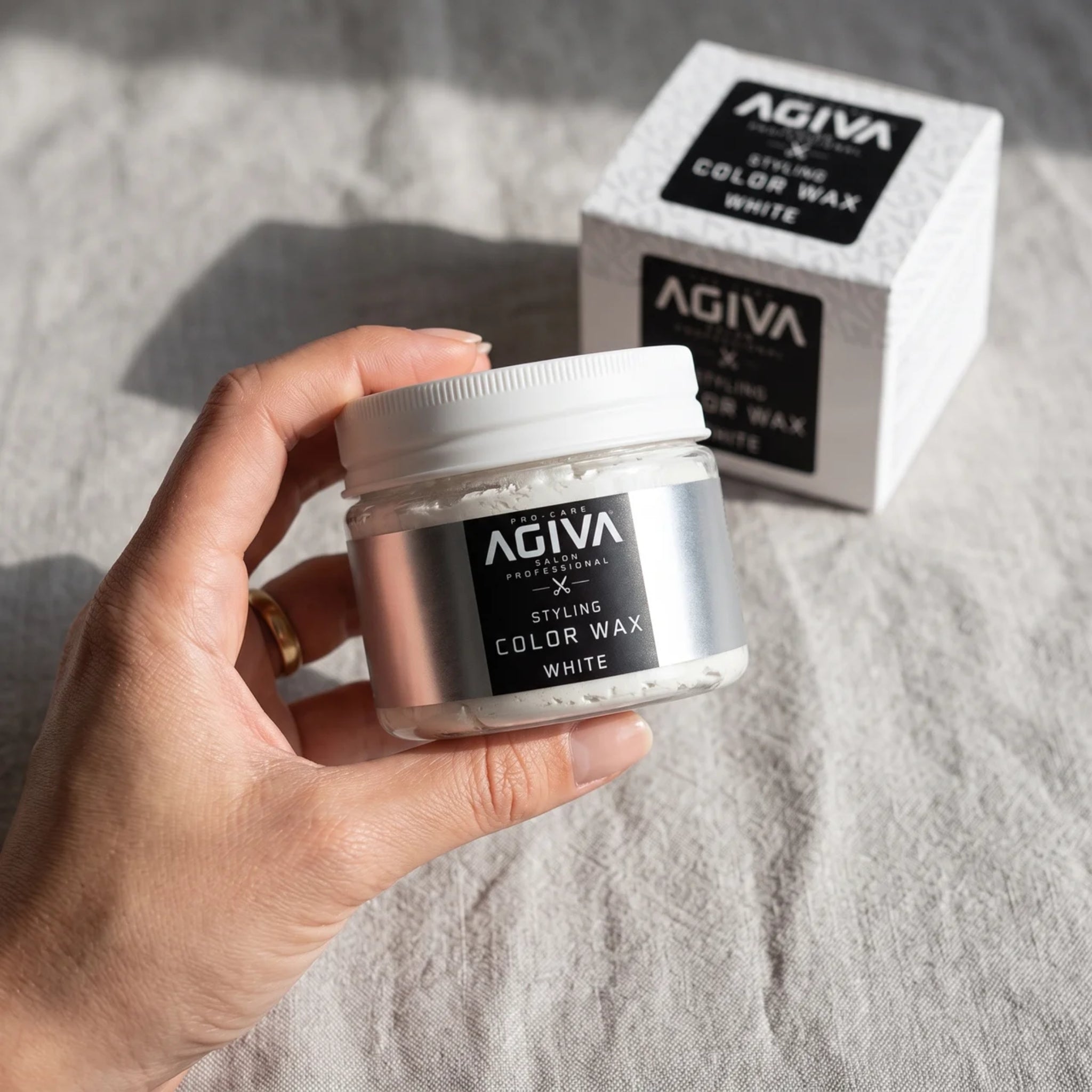 Hånd holder åben dåse med AGIVA Styling Color Wax White, æske med samme produkt ses i baggrunden.