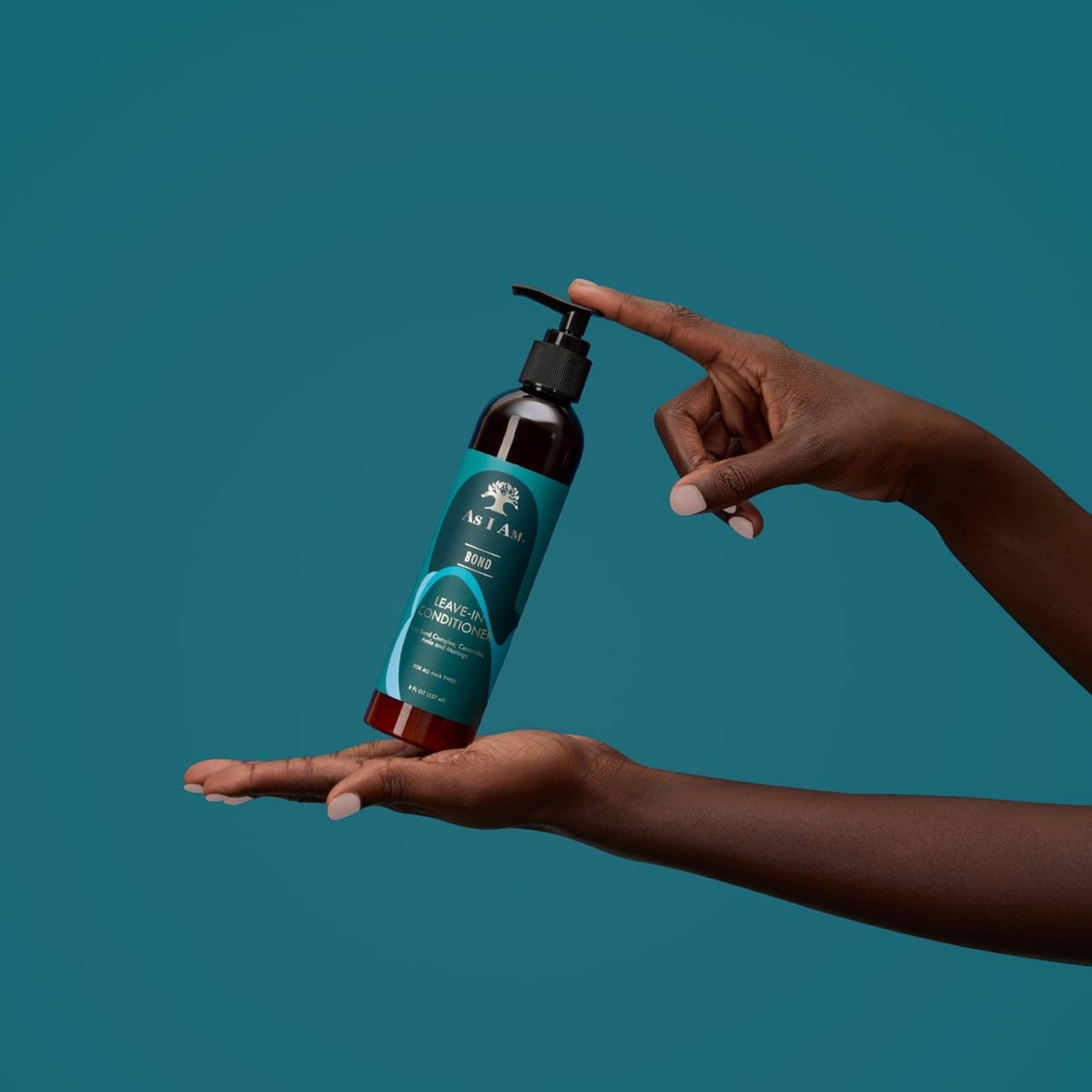 As I Am Bond Leave-In Conditioner flaske holdt mellem to hænder mod petroleumsblå baggrund.