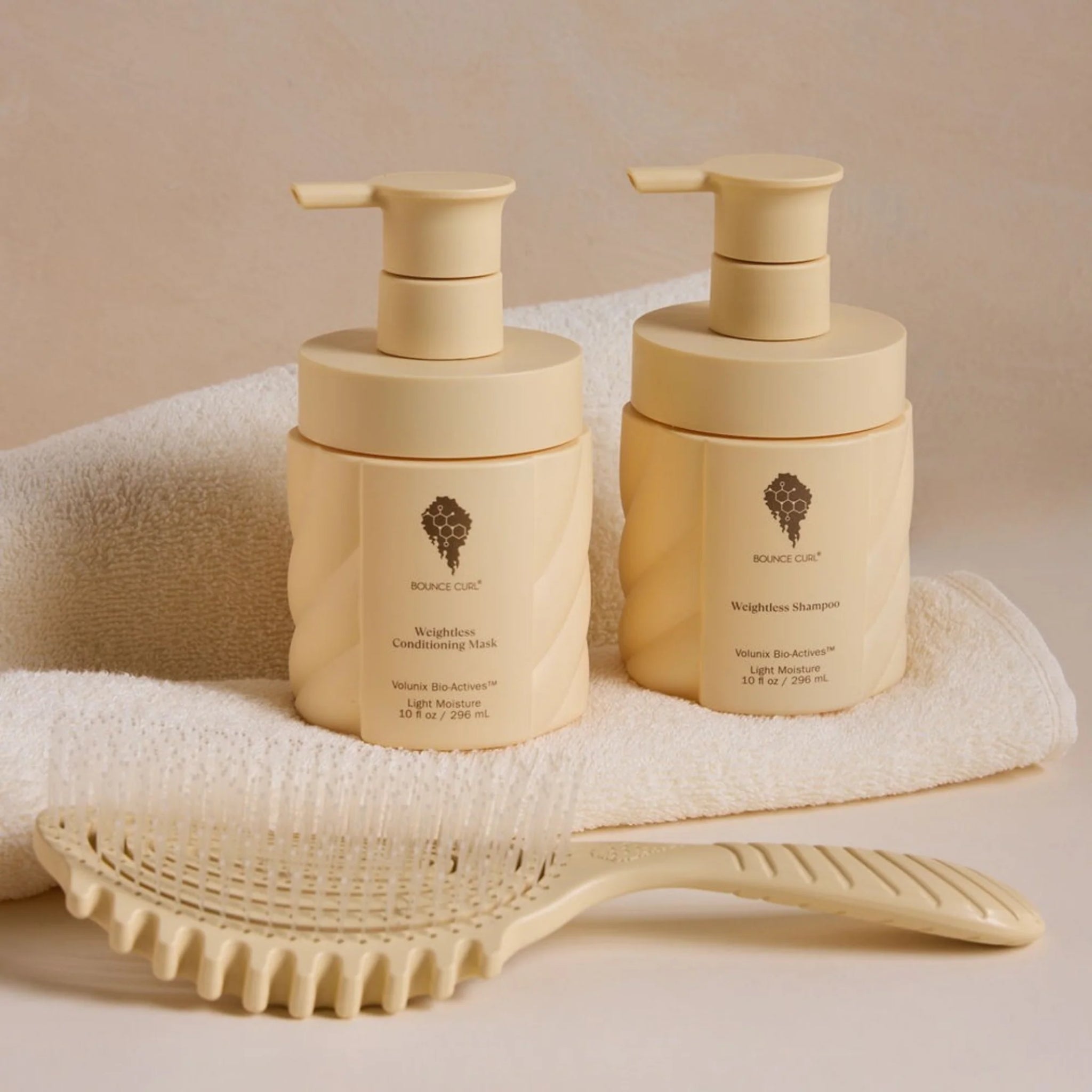 Bounce Curl Weightless Shampoo og Conditioning Mask i cremefarvede flasker med pumpe, placeret på hvidt håndklæde ved siden af hårbørste.