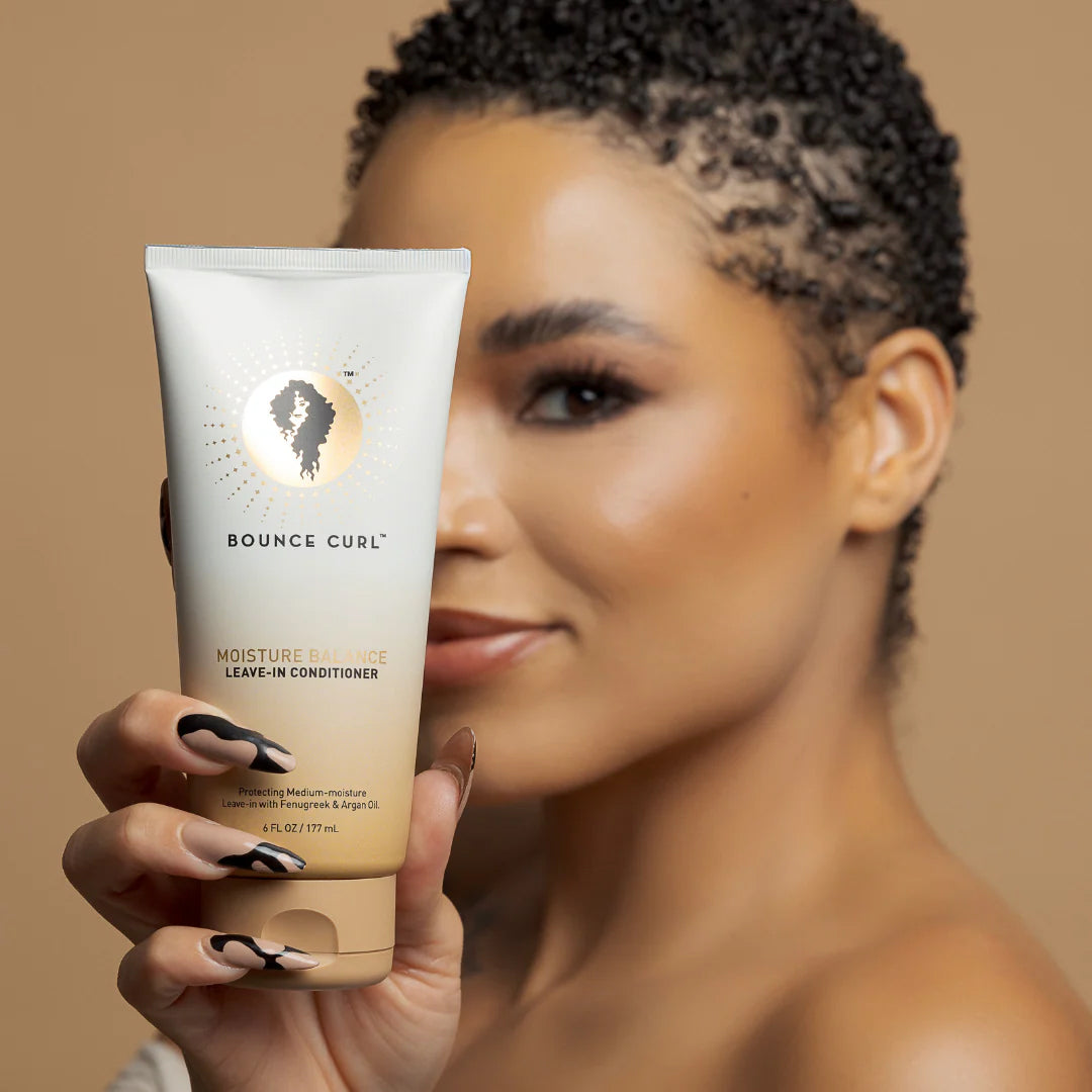 Kvinde med glødende hud holder Bounce Curl Moisture Balance Leave-In Conditioner 177ml, fugtgivende pleje til krøllet hår.