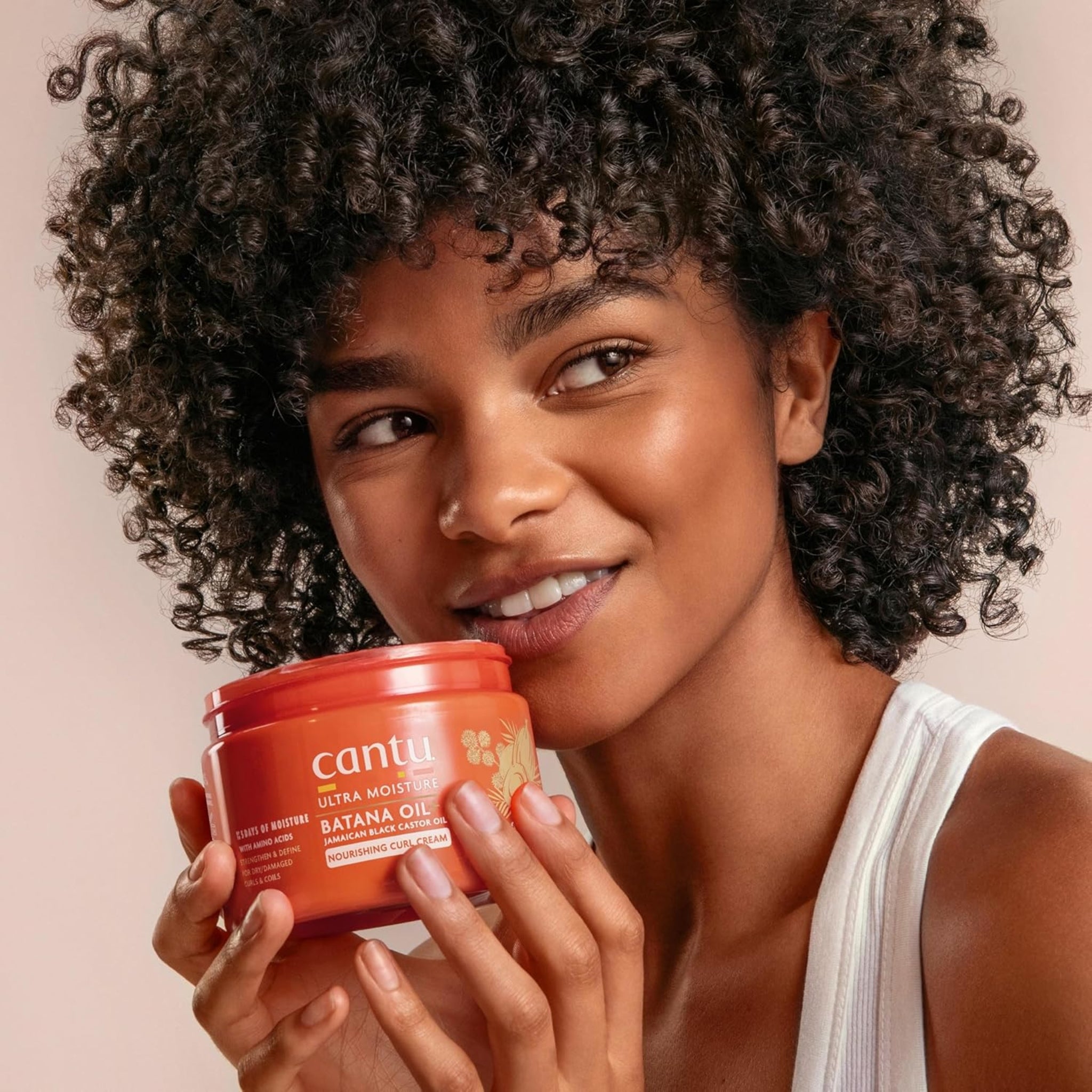 Kvinde med krøllet hår smiler og holder Cantu Ultra Moisture Batana Oil Nourishing Curl Cream op mod kinden.
