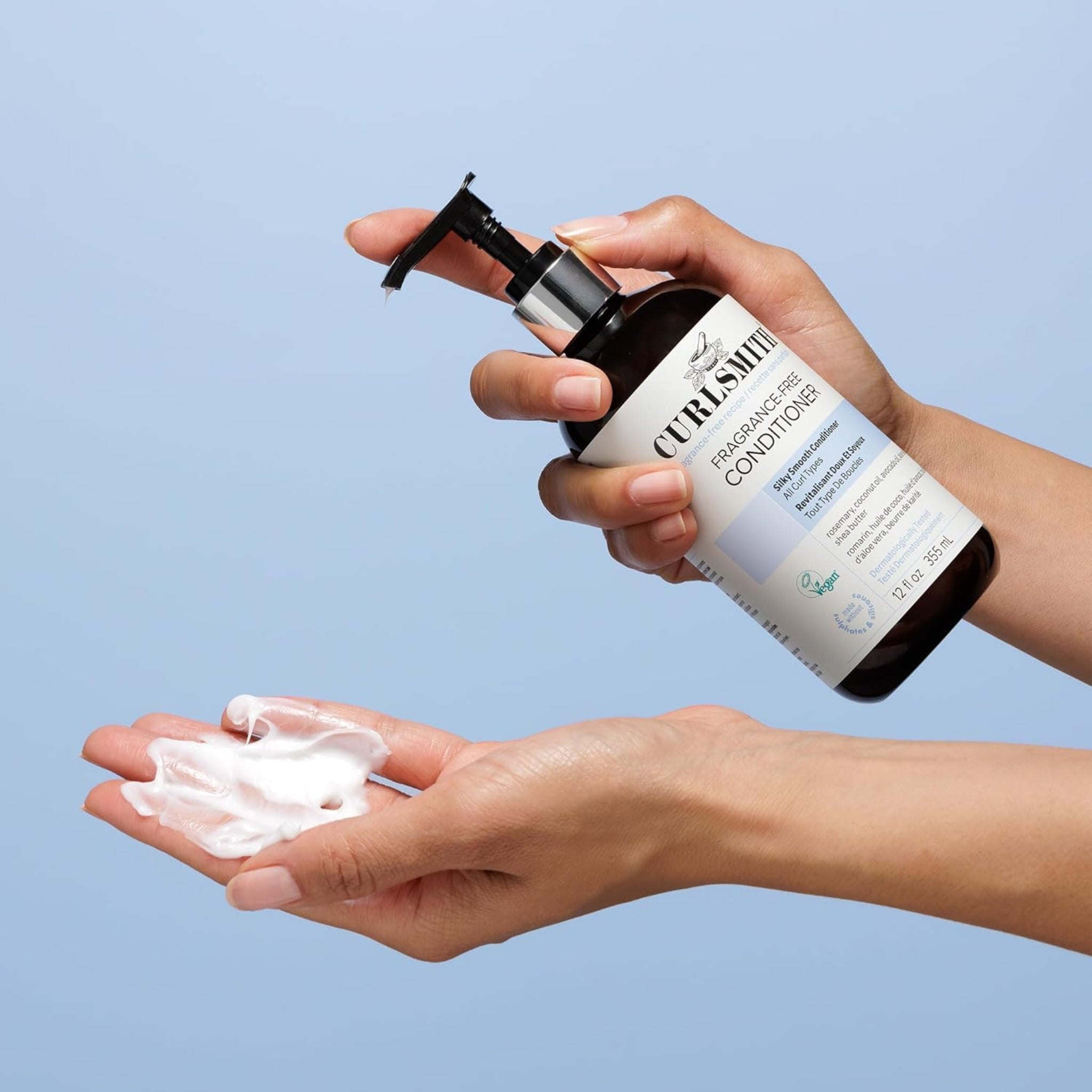 Hånd holder Curlsmith Fragrance-Free Conditioner og pumper hvid creme ud i den anden håndflade mod blå baggrund.