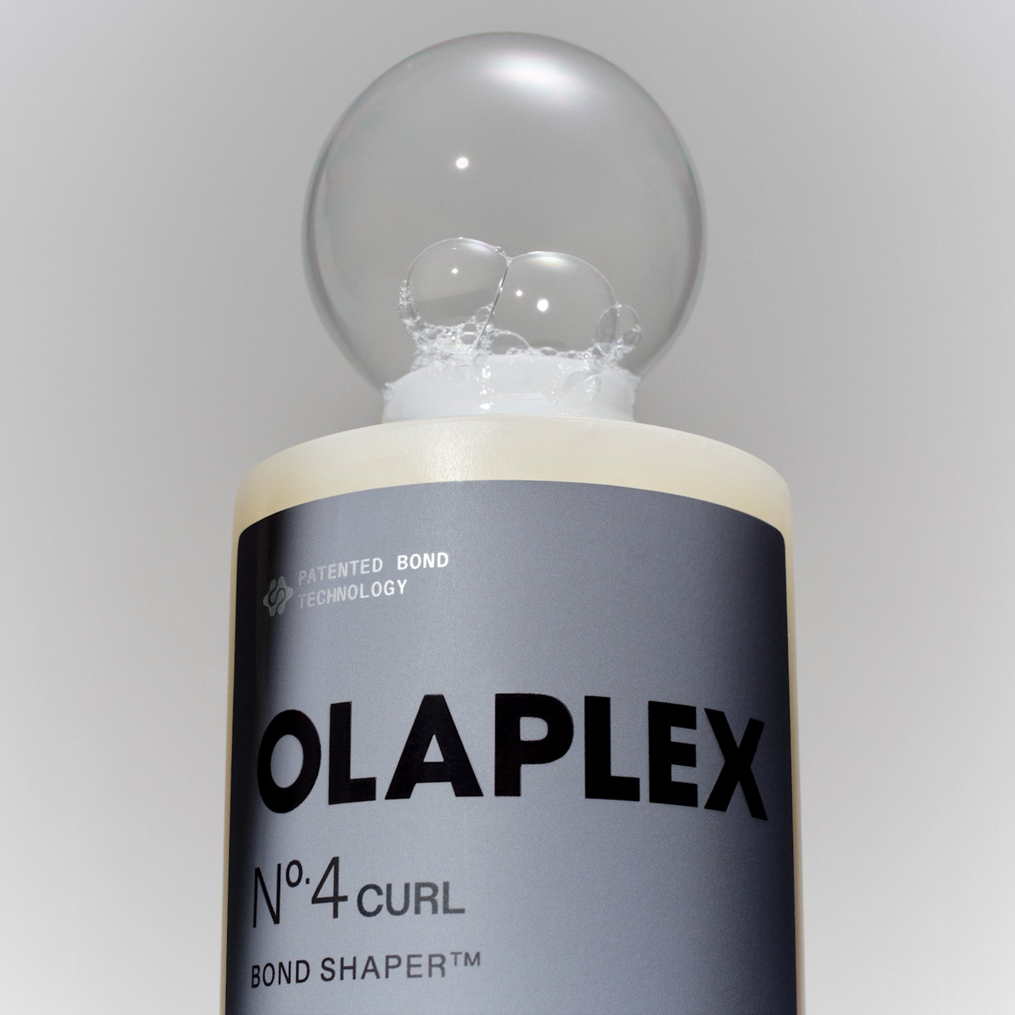 Olaplex Nº.4 Curl Bond Shaper med skum og bobler på toppen mod grå baggrund.