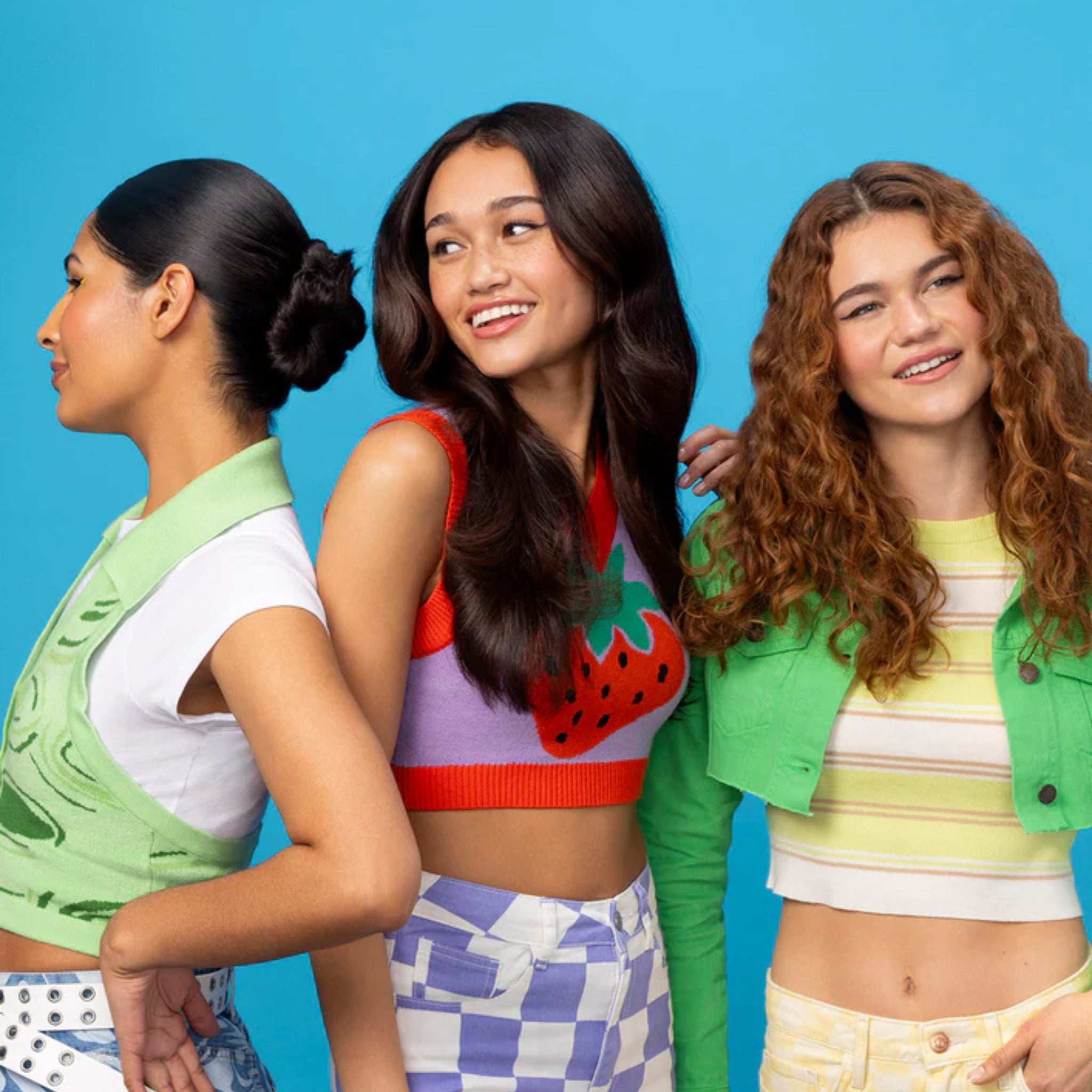 Tre smilende unge kvinder i farverigt sommertøj med jakker og croptops mod blå baggrund.