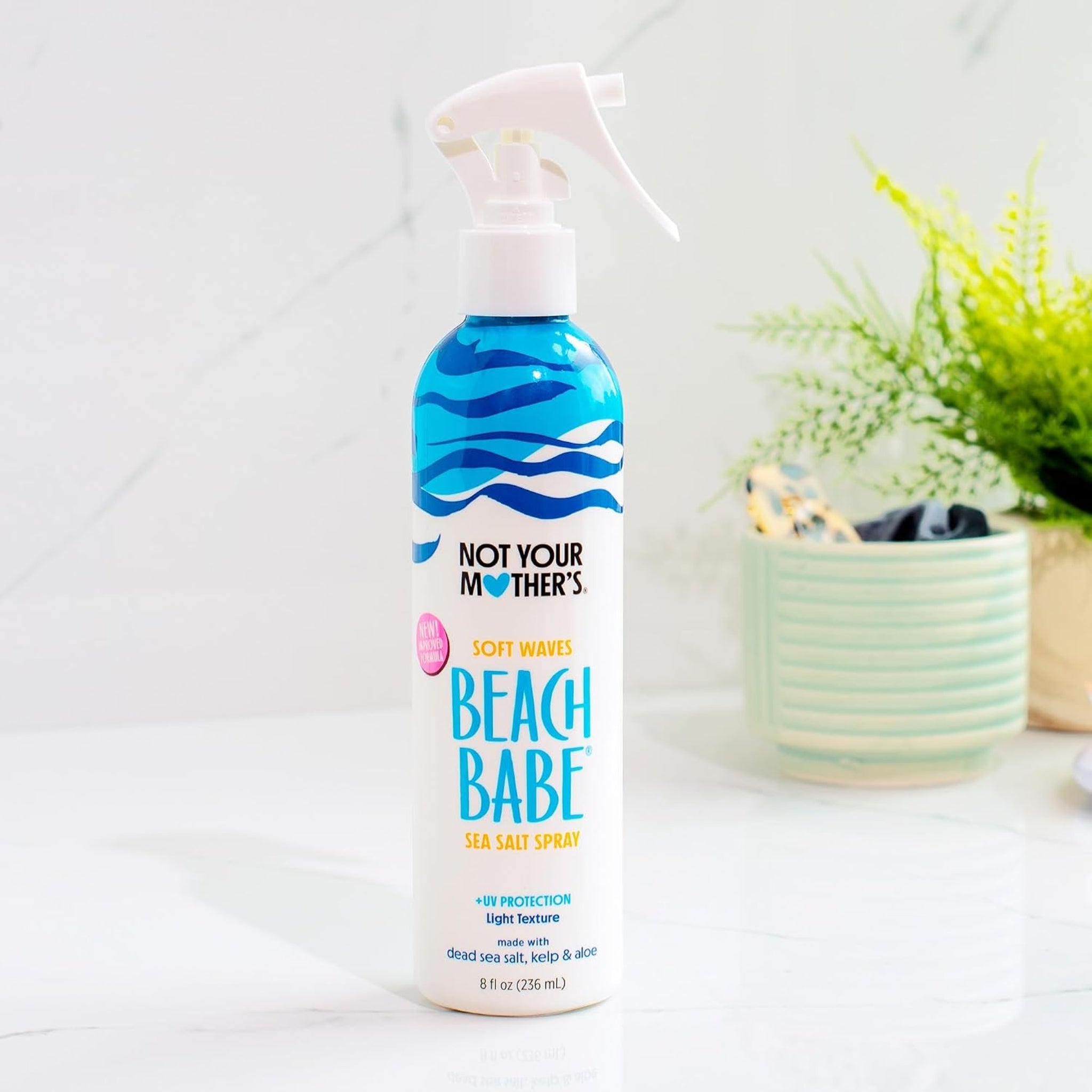 Not Your Mother's Beach Babe sea salt spray i sprøjteflaske på marmorbord ved siden af grøn plante.