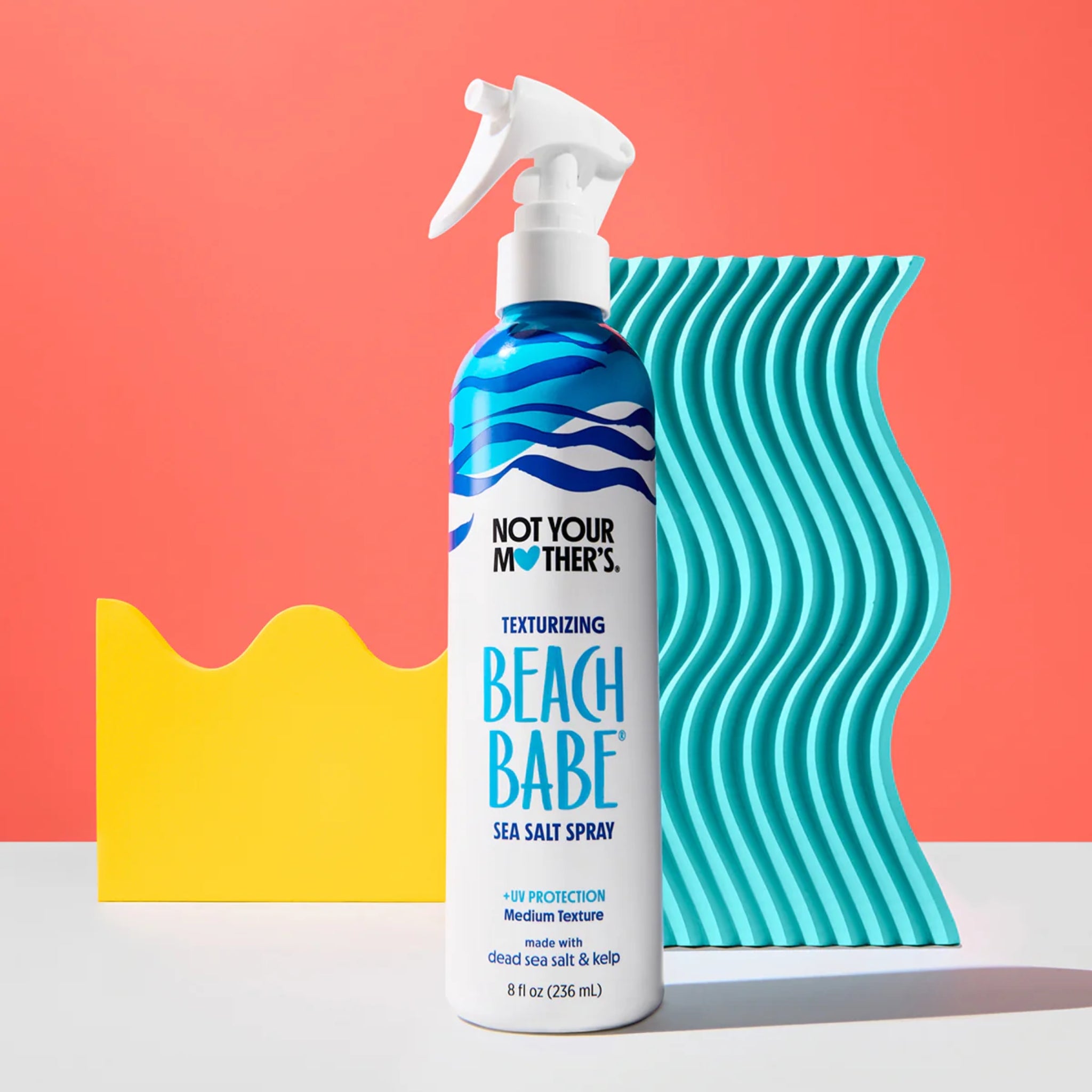 Not Your Mother's Beach Babe sea salt spray foran turkis bølgemønster på koralrød baggrund.