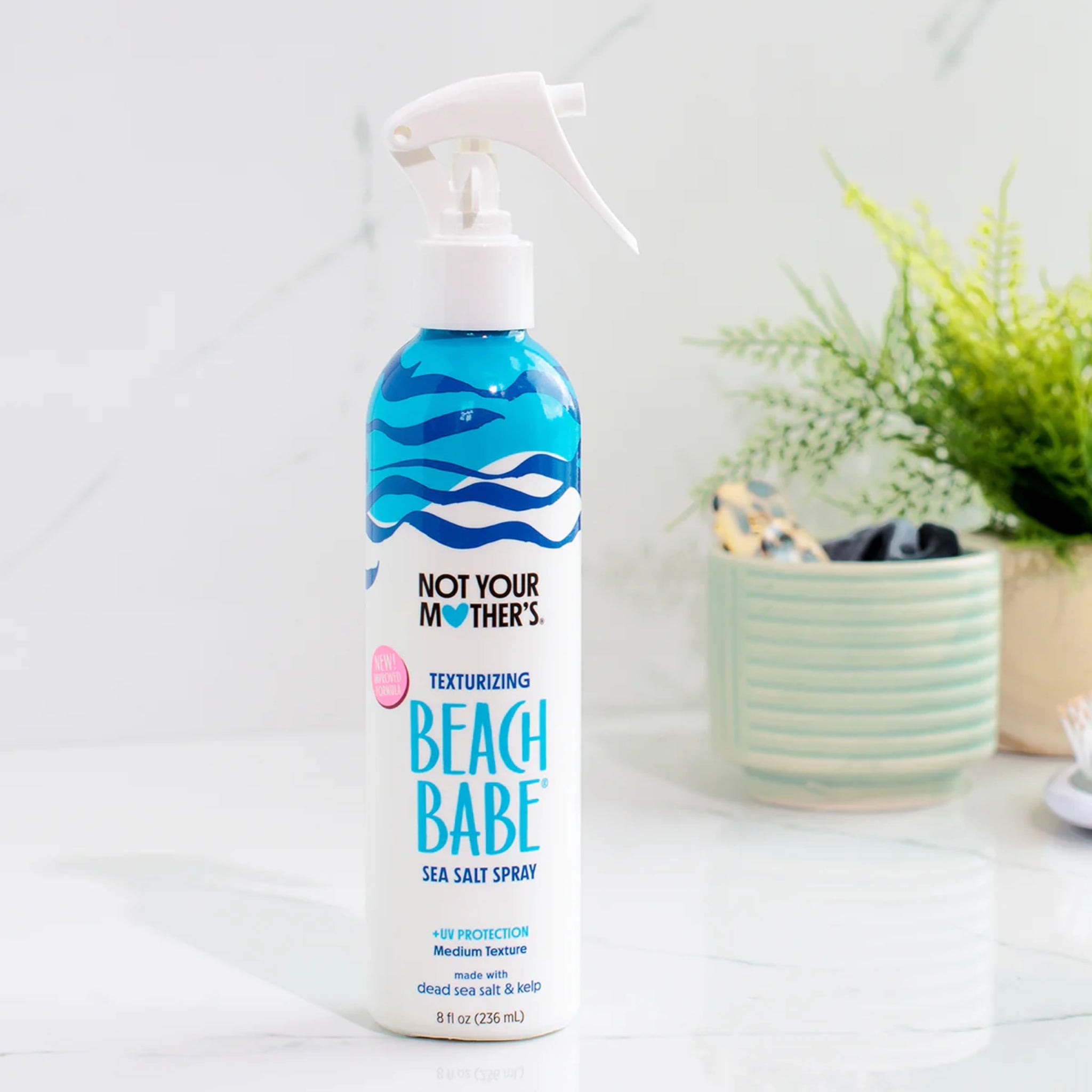 Not Your Mother's Beach Babe sea salt spray i hvid flaske med blå bølger på marmorbord ved grøn plante.