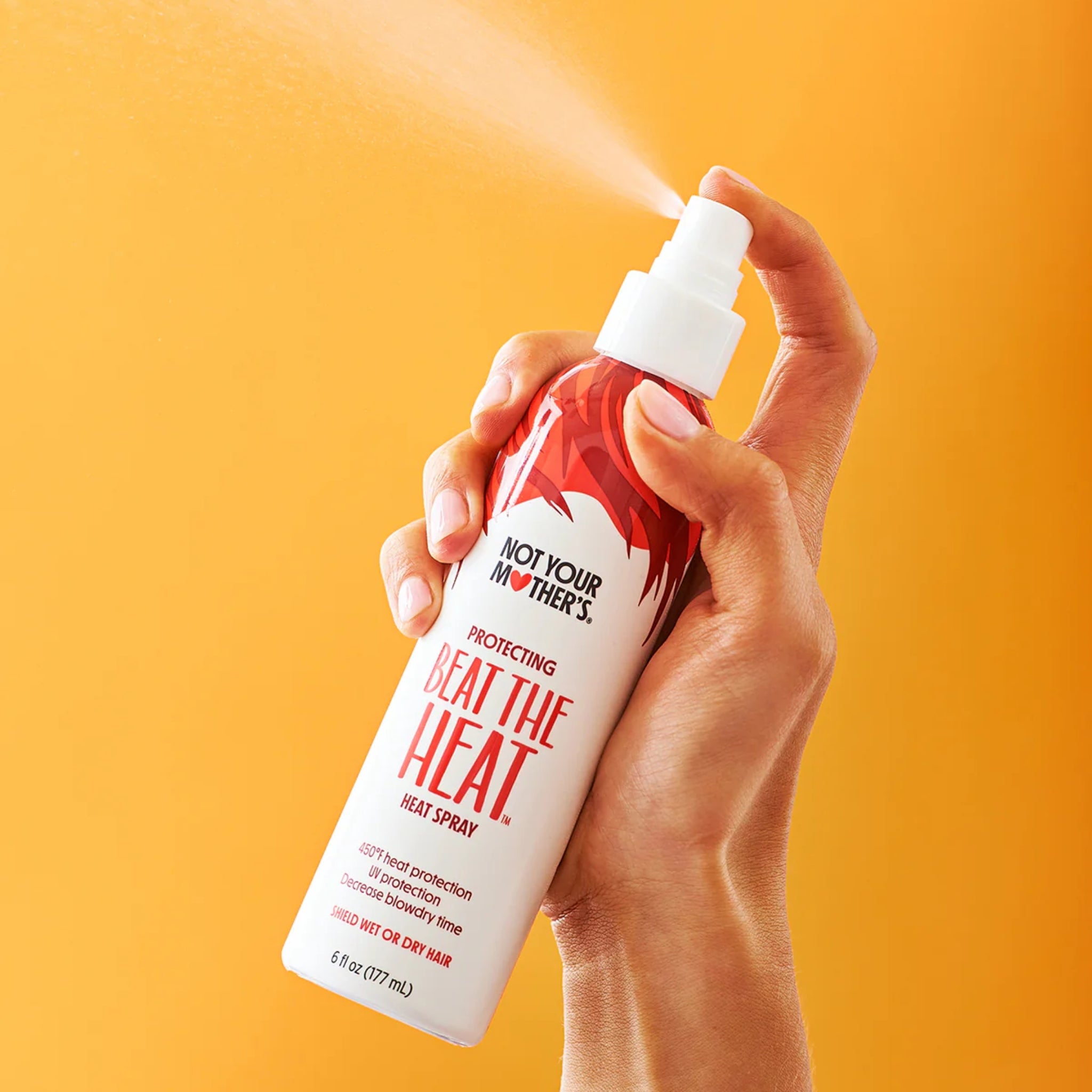 Hånd der sprayer Not Your Mother's Beat The Heat varmebeskyttende hårspray mod orange baggrund.
