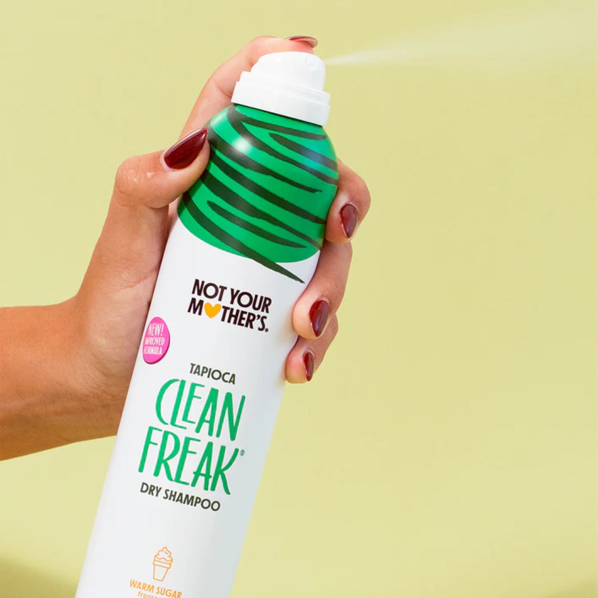 Hånd holder Not Your Mother's Clean Freak tørshampoo i grøn dåse, der sprøjtes mod grøn baggrund.