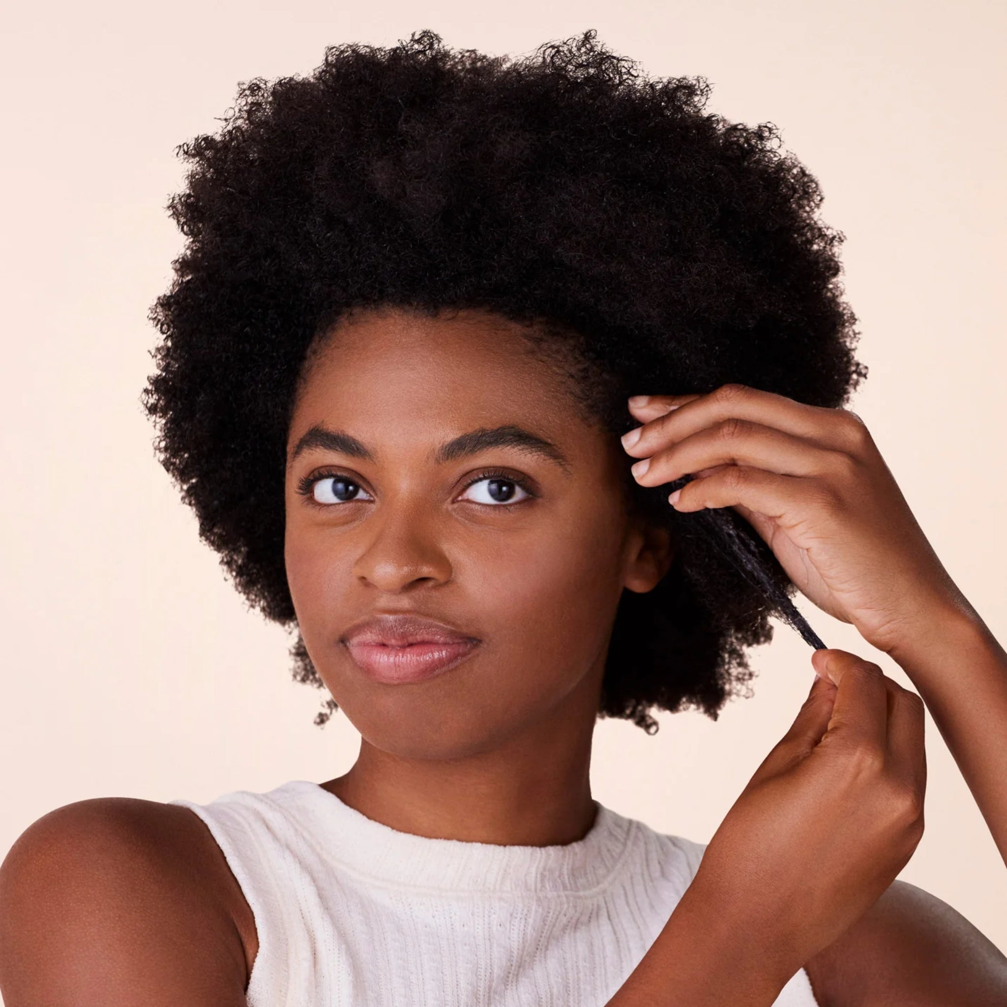 Kvinde med naturligt afrohår sektionerer sit krøllede hår med fingrene mod en beige baggrund.