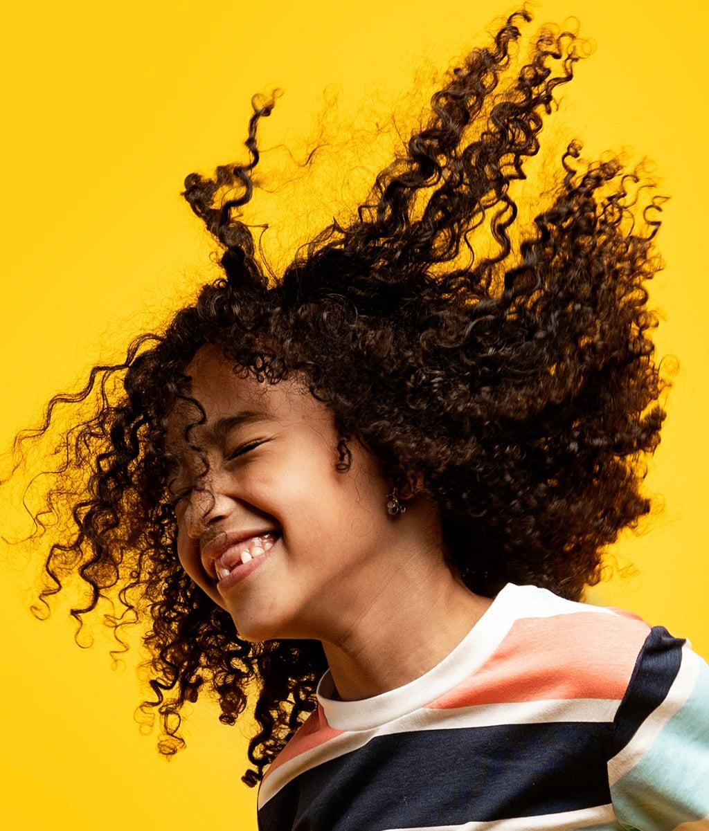 Barn med glade, definerede krøller efter brug af Not Your Mother's Kids Curly Care Cream 237ml mod gul baggrund.