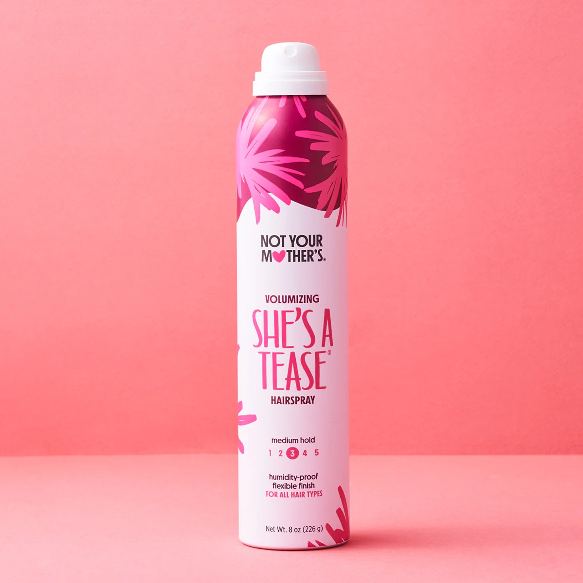 Not Your Mother's She's A Tease volumizing hairspray i lyserød dåse på koralfarvet baggrund.