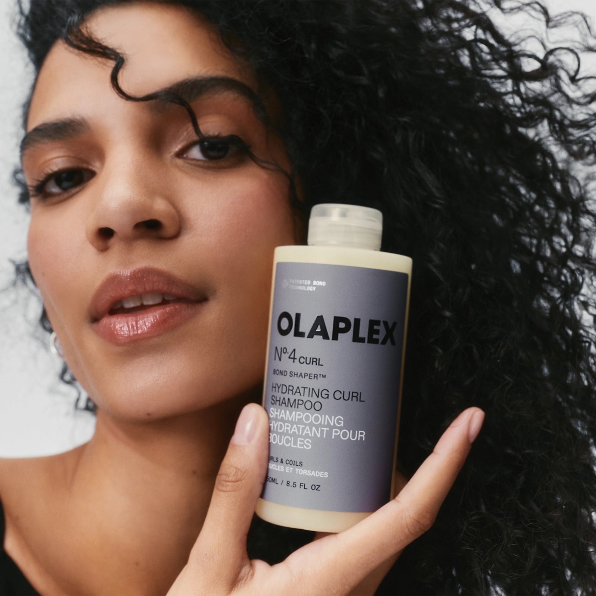 Kvinde med krøllet hår holder Olaplex Nº.4 Curl Hydrating Curl Shampoo til lokker og krøller.