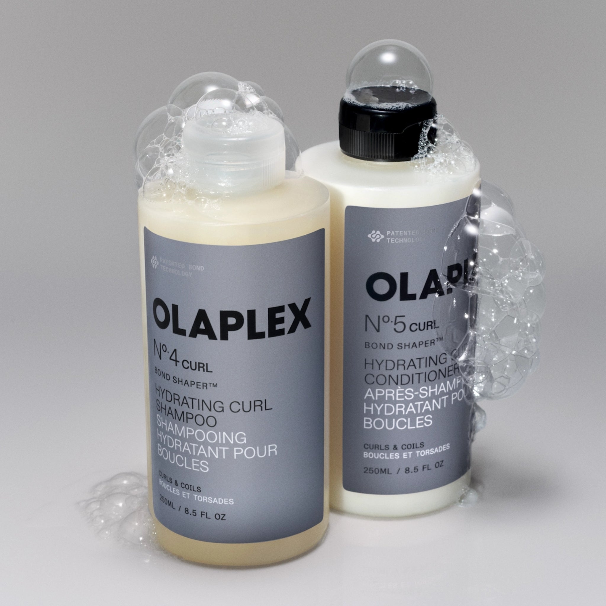 Olaplex Nº4 Curl shampoo og Nº5 Curl conditioner i 250 ml flasker omgivet af sæbeskum.