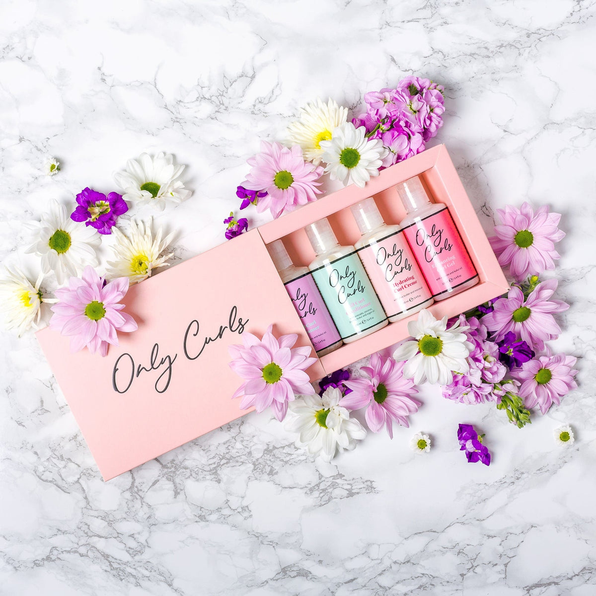 Only Curls gaveæske med fire hårplejeprodukter i pink emballage, omgivet af lilla og hvide blomster på marmorbord.