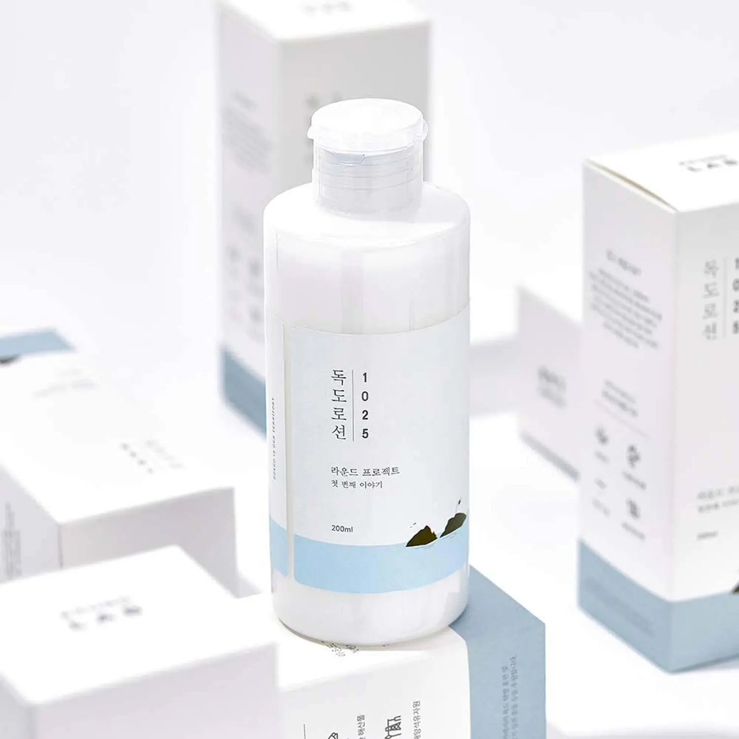 Round Lab 1025 Dokdo Lotion 200ml i hvid flaske med blå etiket, placeret blandt produktæsker i lys baggrund.
