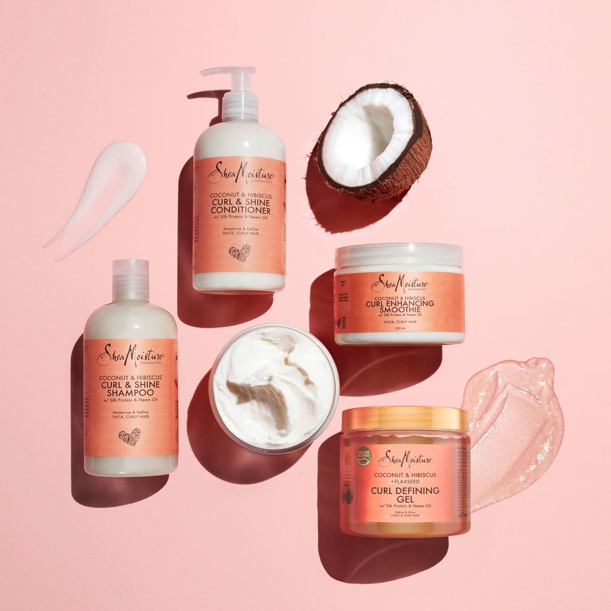 SheaMoisture Coconut & Hibiscus hårplejeserie med shampoo, balsam, curl smoothie og curl gel ved halveret kokosnød på lyserød baggrund.