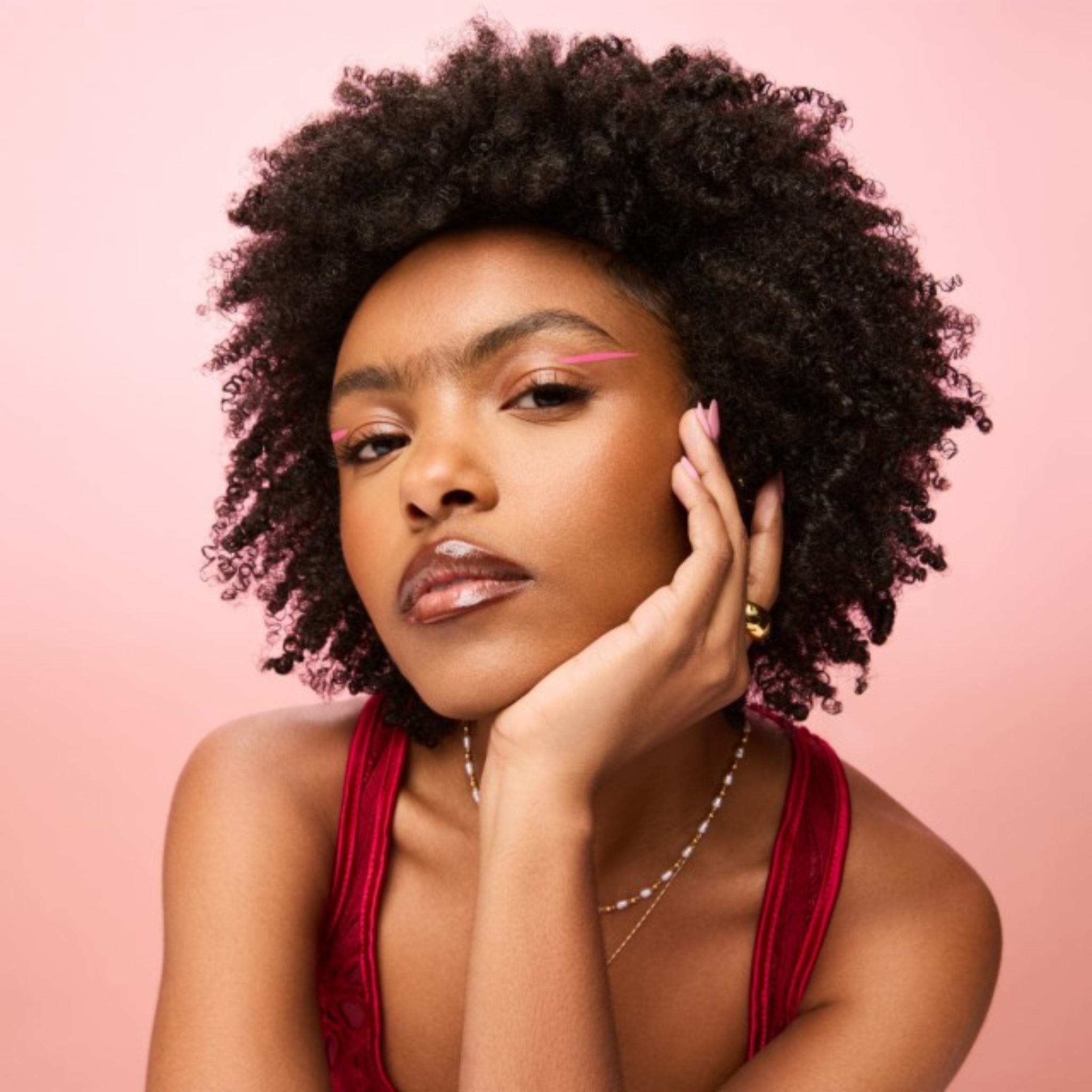 Kvinde med naturligt afro-hår og pink eyeliner hviler ansigtet i hånden mod lyserød baggrund.